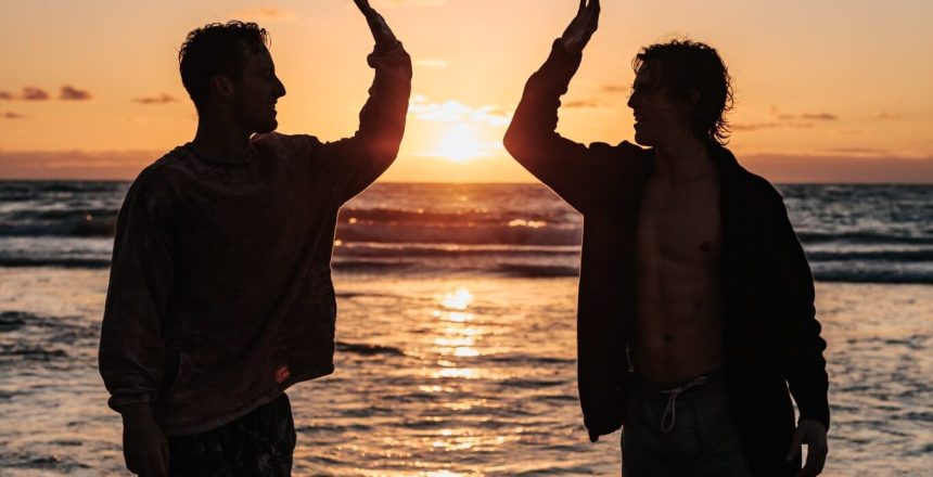 Silhouette of two people high fiving on a beach with a sunrise between them.